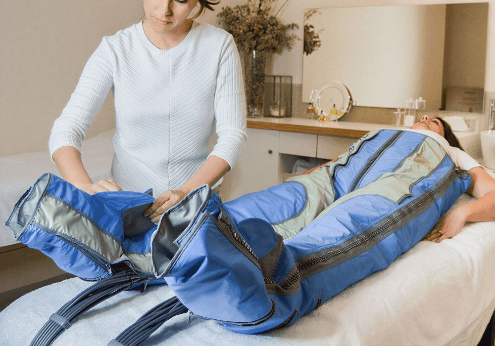A serene spa or wellness center scene featuring a client lying comfortably on a white treatment bed, fully encased in a blue and gray inflatable compression therapy suit with visible zippers and segmented chambers designed for body contouring or lymphatic drainage. A female therapist dressed in a white ribbed long-sleeve top is gently adjusting the leg portion of the suit, focusing on proper fit and alignment. The background includes a softly lit, minimalist interior with a wooden countertop, decorative dried floral arrangements in a vase, a round mirror, and neatly arranged skincare bottles, creating a calming, professional aesthetic suitable for promoting premium beauty or body treatments.
